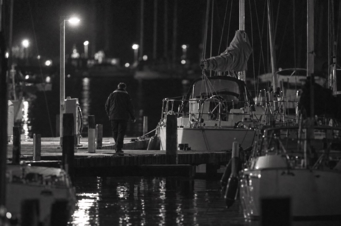 Long-lens night photo of a man in a club jacket walking along a pier